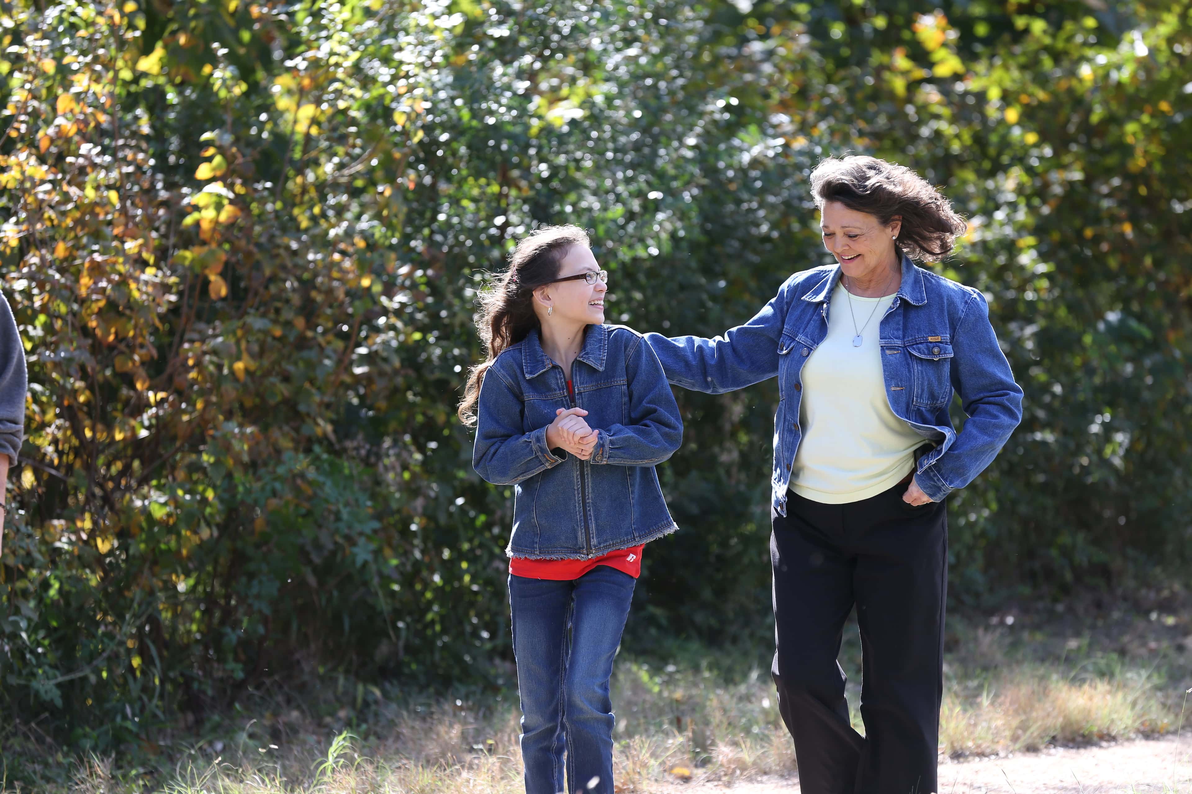 A girl in a denim jacket walking outdoors, smiling beside a woman.